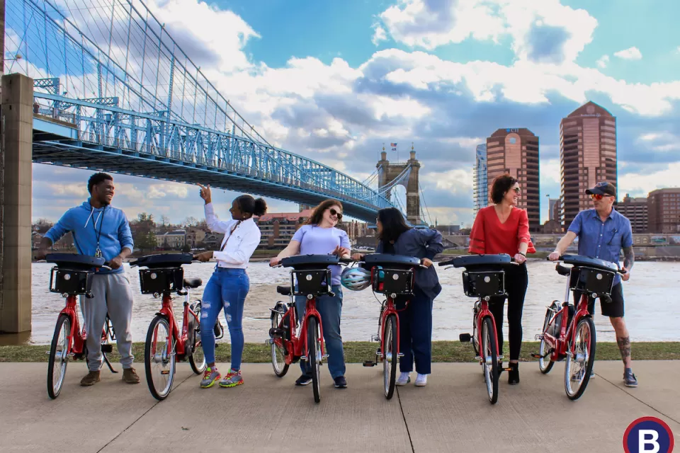 Six people posing with Red Bike bike share bikes with bridge and river in background.